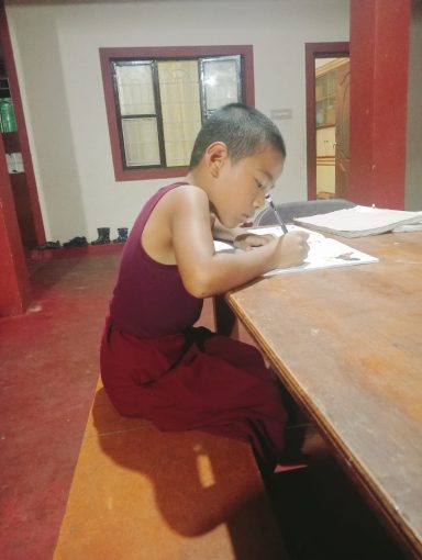 Niño monje escribiendo en un cuaderno sobre una mesa en un ambiente tranquilo.