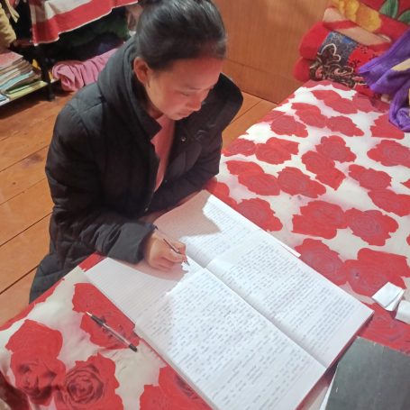 Mujer sentada escribiendo en un cuaderno sobre una mesa con un mantel rojo.