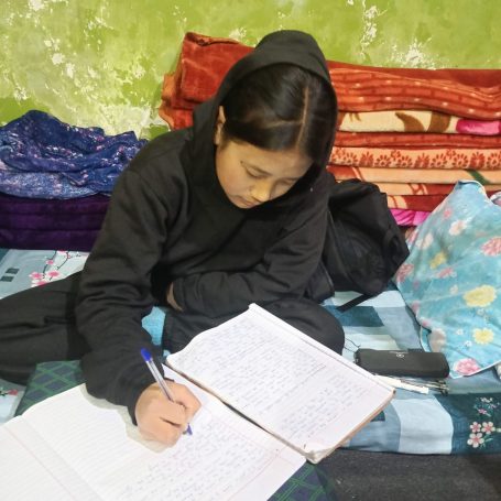 Niña con ropa negra, escribiendo en un cuaderno sobre una cama con mantas.